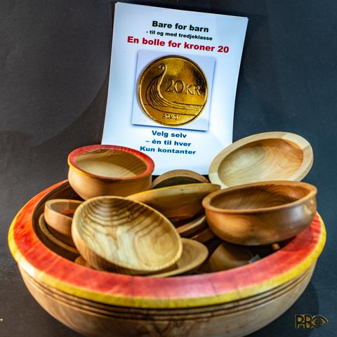 In a huge wooden bowl made of birch with red decoration around the opening is a pile of smaller turned bowls and asjets. A poster stands in the background with a large twenty kroner coin in the middle with text above and below that says "Only for children - up to and including third grade - A bowl for kroner 20 - Choose yourself - one for each - Cash only