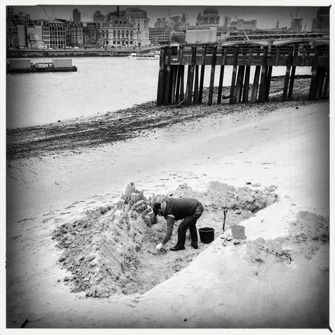 A b/w photo of a man creating a sand castle on the South Bank of the River Thames in London, UK.