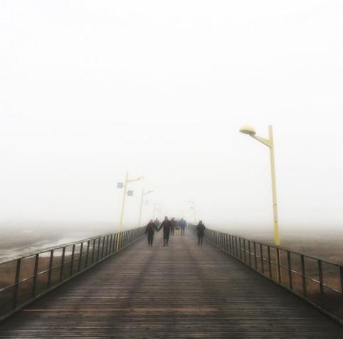 Several people walk the pier of St. Peter-Ording on a grey and foggy day.
