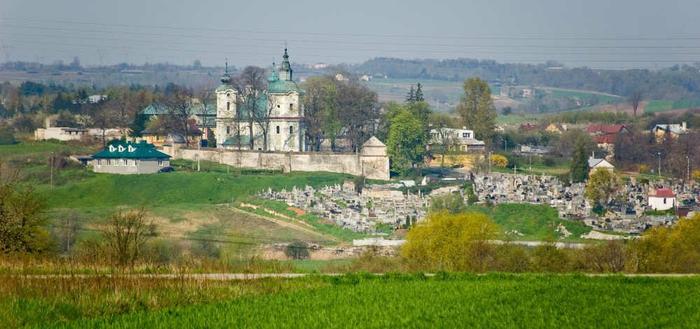 Panorama z lewej strony w oddali kościół. Od środka kadru do końca prawej strony cmentarz. Na samym dole pole z trawami. W górnym kadrze niebieskie niebo.