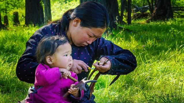A woman and child sit among green grasses. Mother is intently carving a reindeer antler with a screwdriver while the young child pretends to do likewise with a small stick and a small antler.