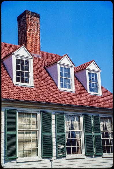 A portion of the top floor and roof of a wood-framed building. The white building has green shutters and a reddish shake roof. Across the top floor, three double-hung windows are visible (the building extends out-of-frame to the left). The left-most window has blinds inside, while the other two have parted curtains. Three smaller dormer windows protrude from the roof. A square brick chimney extends from the peak of the roof. Beyond, the sky is a deep and cloudless blue.