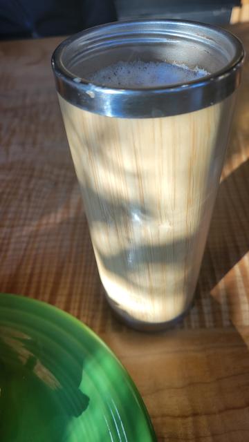 Photo of a tall wooden go cup with the foam of a mocha showing at the top, sitting on a wooden table by a bright green saucer. Sunlight is streaming across from the left, casting shadows to the right.