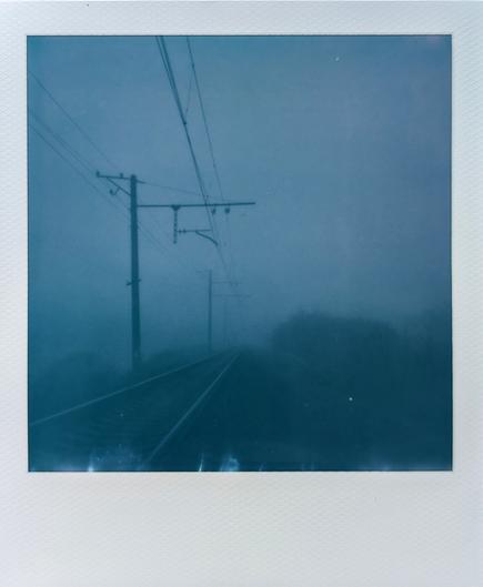 Instant Polaroid photo.
A railroad track stretching from the lower left corner into the distance and disappearing into the fog.
Along the track are poles with stretched wires.
Everything around is covered with thick fog. The entire image is in blue and green colors. In general, it creates a mystical and enigmatic atmosphere
