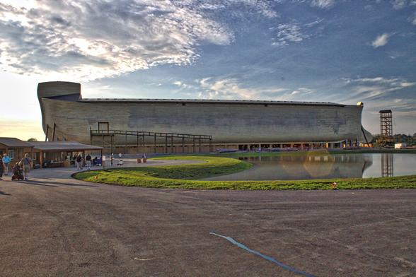 A photo of the Ark Encounter, a purported replica of Noah's Ark but built with ferro-concrete. The sky is partly cloudy with the sun low in the sky behind the Ark. In the left foreground, a few people are walking towards the ark on a paved pathway past the gift shop. On the right is an artificial pond.