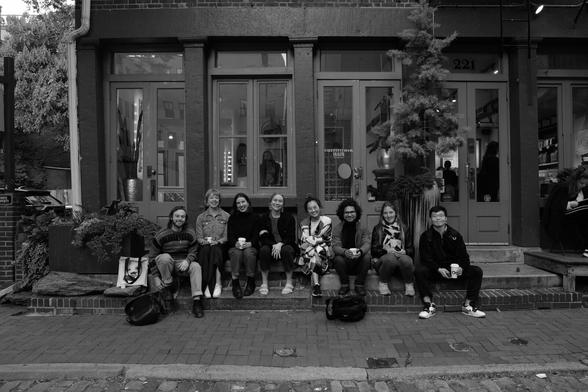 A black-and-white picture of a group of 8 people sitting on the sidewalk of Philadelphia old town
