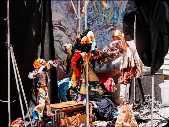 A color photograph showing behind the scenes at a puppet theater set up outdoors. Several marionettes are hanging in front of a painted backdrop showing leaping dolphins, the black fabric curtain, and a steamer trunk. The detailed marionettes include an Emmet Kelly-esque clown, a witch in green on a broomstick, and a Brunhilda-like figure. Beyond behind that, the shaggy form of a dog marionette is just visible. Cables, likely audio, containers and bags as it on the grass below.
