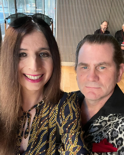 Left: woman wearing gold and black pattern.
Right: man wearing silver leopard-print outfit.
Inside a small concert hall with a window looking out towards a busy city street.