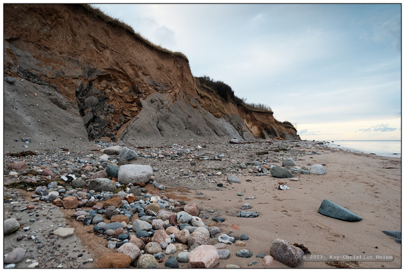 Weitwinkelaufnahme des Steilufers an der Ostsee, im Vordergrund links sind tiefe Ausspülungen und schichtartige Strukturen im blankgespülten Lehmboden der Abbruchkante zu sehen. Rechts im Hintergrund die ruhige Ostsee hinter einem steinigen, von der Sturmflut glattgehobelten Strand.