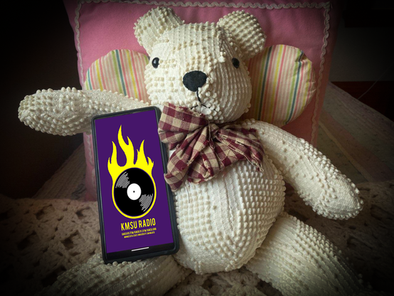 Photo of a fabric toy bear holding a smartphone with the KMSU RADIO app on the screen.