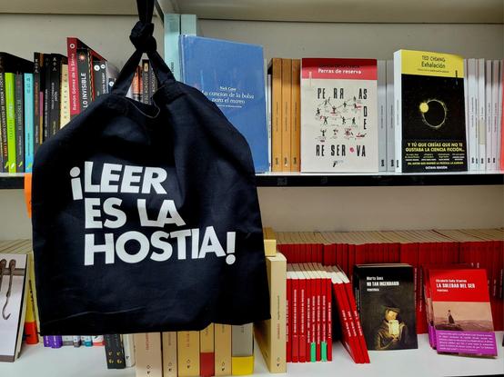 Se ven varios estantes de la caseta 14 de la FLS con libros variados. Colgada del estante superior está la bolsa negra de ¡Hostia un libro!, que reza en letras blancas, con el diseño de Puño, "¡LEER ES LA HOSTIA!"