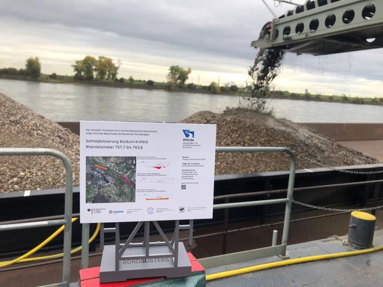 Eine Schautafel gibt Informationen zum Projekt. Im Hintergrund das Förderband, das Rheinkies in eine parallel liegende Schute transportiert.