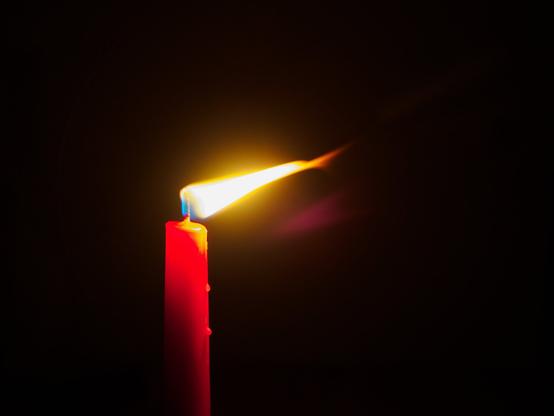 A lit red candle. The background is dark, nearly black. There is wind coming from the left and the flame is being pushed to the right.