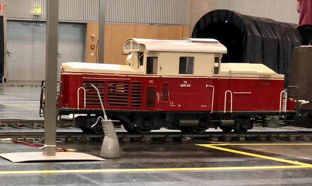 Ein elektrisch betriebenes Modell einer 2091.03 in historischer Lackierung. (Unten dunkelrot und oben beige.) Der Zug halt an einen Ausfahrsignal dessen Mast an der Zugspitze zu erkennen ist. Neben der Lok steht ein Ölkännchen im Maßstab 1:1.