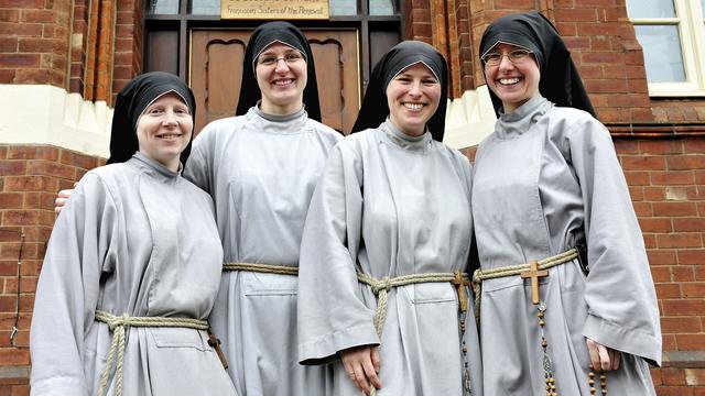 A group of four smiling nuns in front of a big door. They're wearing grey habits and black headscarves