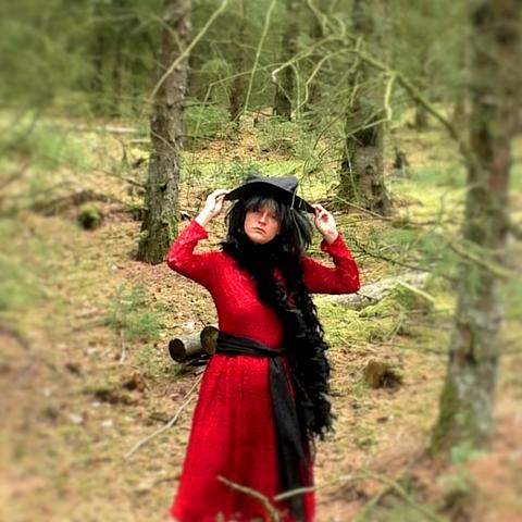 Halloweenkostüm: Eine Hexe in rotem Kleid, schwarzem Gürtel, schwarzem Schal und schwarzem Spitzhut, den sie gegen den Wind festhält.