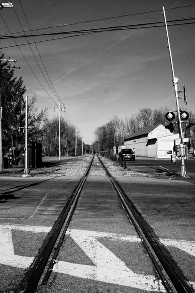 A black and white photograph, shot vertically, taken from the middle of a railroad crossing looking down the tracks. The paved road the rails are embedded in is visible in the foreground, a crosswalk running right to left across the tracks.

A line of utility poles is visible on the left side of the tracks, and a commercial building with a truck sits to the right past where the road ends and gravel ballast and railroad ties resume around and between the tracks. Bare trees are visible on either side in the distance. An open railroad crossing gate and a set of warning lights are visible nearby on the right. A think layer of clouds is slightly visible, along with an aircraft contrail.