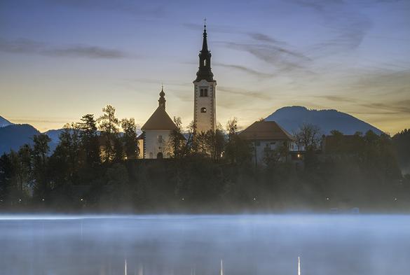 Early morning view of Lake Bled island in Slovenia with the spires of the church and other buildings silhouettes against the dawn sky. Fog rising from the lake in the foreground.