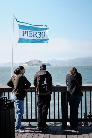 A group of people looking out at Alcatraz, standing underneath a flag reading "PIER 39"