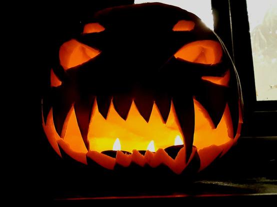 A scary Halloween pumpkin sitting on a countertop with fire-red light emanating from it. It has a big sharp-toothed grin and gleeful eyes. It appears to be laughing malevolently.