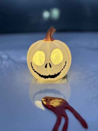 A toy pumpkin, with eye holes and drawn on nose holes and a long, twisted mouth glows yellow on a white car hood with a red splotch of blood.