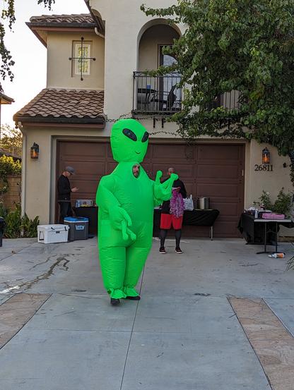 A man in an inflatable green alien suit with glowing eyes stands in a drive way before a big trick or treat caravan in the suburbs.