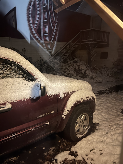 A jeep liberty sites in a driveway. Both the ground and the jeep are blanketed in an inch of snow.