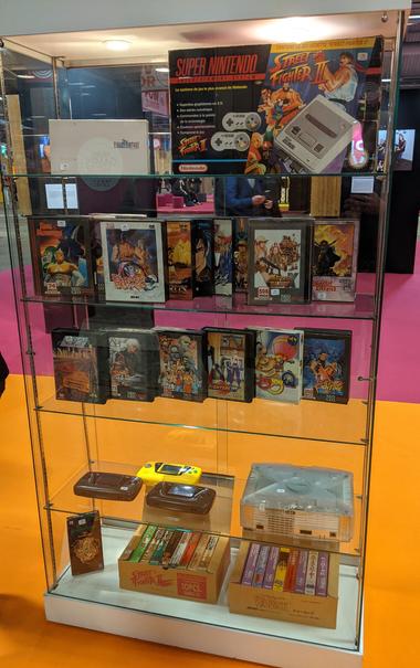 Vitrine avec objets mis aux enchères : Super Nintendo Street Fighter II dans sa boîte, consoles Sega Game Gear, Xbox, jeux Neo-Geo, etc.