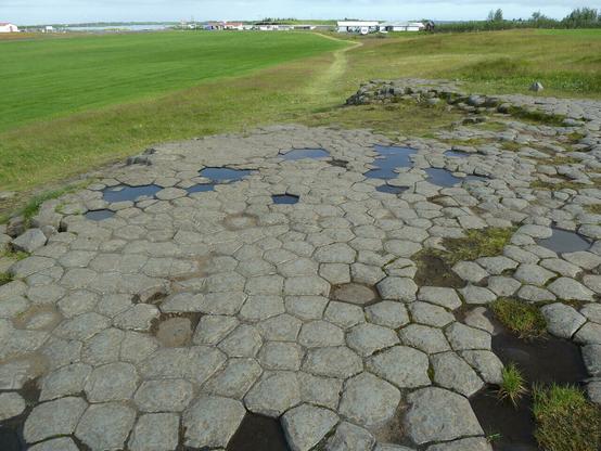 Im Vordergrund graue Polygone mit dunkleren Rändern auf dem Boden, dahinter Wiesen, in der Mitte ein Pfad, ganz hinten Bebauung. Einige Polygone scheinen zu fehlen oder abgesenkt zu sein; dort steht Wasser.