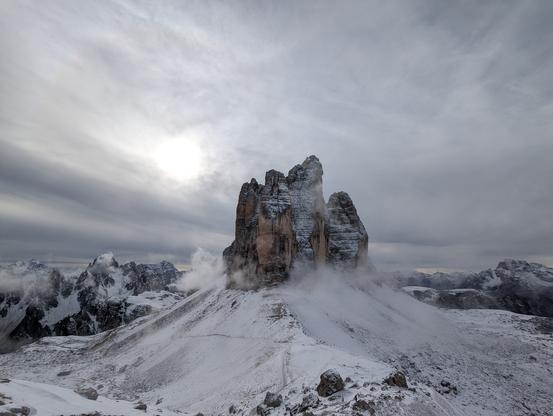 Winterliche Aufnahme der Drei Zinnen von Norden aus. Der Himmel ist vergangen mit Wolken, die Sonnen scheint links der Drei Zinnen leicht durch. Vor den Bergen zieht eine Wolke wie eine Nebelschwade durch