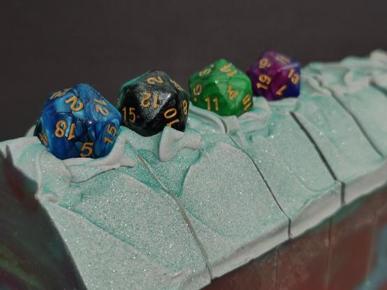 A sparkly green mounded soap top with one blue, one black, one green and one purple D20 on top