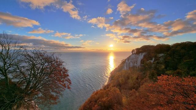 Wir stehen über dem Kreidefelsen Königsstuhl, also über 120 m über der Ostsee und schauen auf das Meer.
Rechts von uns sehen wir im herbstlichen Buchenwald den Kreidefelsen unter der Victoriasicht.
Zwischen uns und der Victoriasicht fällt die bewaldete Küste sanft ins Meer und wir sehen ein kleines Stück Strand. Steinstrand.
Das Highlight ist jedoch die direkt über dem Horizont aufgehende Sonne. Sie entsteigt der Ostsee und spiegelt sich in einem Streifen über das Meer bis zum Strand. Das warme Licht taucht alles in einem goldenen Schein.