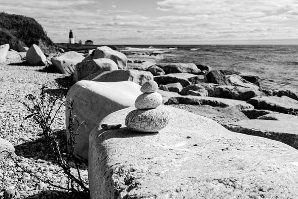 A black and white photograph taken along a rocky shoreline looking towards a lighthouse in the distance. A line of large rocks sits as a divider between the land and water. On the nearest one, a small pile of three smooth, oblong rocks has been built, stacked largest to smallest. A coastal plant, withered, rises from the geound below or behind the large rock.