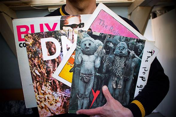 Person with a black sweatshirt with yellow details on the cuffs holding four record albums in his hands by The Contortions, DNA, The Raincoats and The Pop Group.