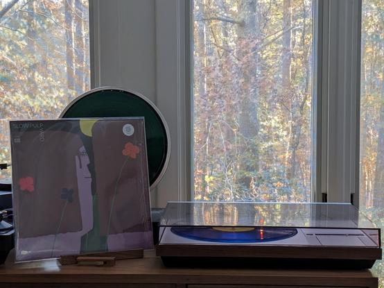 Slow Pulp - Yard LP cover.

A cartoon of two people, flowers behind them, looking out onto a yellow sphere that might be the sun. Who can tell.

The transparent blue LP plays on a vintage Bang & Olufsen Beogram 4000 linear-tracking turntable to the right. The dustcover is closed.