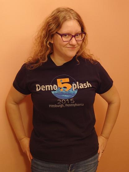 A woman standing with her hands on her hips wearing a Demosplash 2015 T-shirt, with the front design facing the camera.