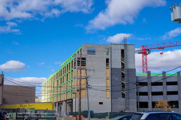 Uneven sunlight broken by clouds lights up the bright green grid of insulation on the wall of a building under construction. The window holes are filled with chipboard panels, and the walls of the building re various shades of grey. A yellow truck is angled up from a loading dock at the left, and cables arc away from two utility poles in front of the building. To the left, a bright red construction crane rises above another bright-green strip of insulation across the top of a dark grey parking structure. Above, a blue sky with puffy clouds.