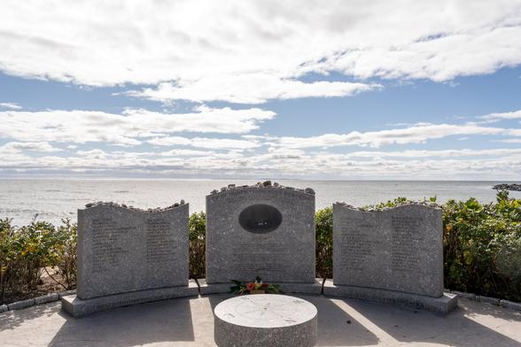 A color photograph of a memorial taken on a bright day with some clouds in the sky. In the foreground, backed by some greenery, three headstone shaped memorial stones stand in a curved line, the center one slightly taller. The left and right stones have swooping tops, evoking sea swells, the center stone has a curved top, and an inset plaque depicting a small sailing vessel.

The center stone reads “POINT JUDITH FISHERMAN’S MEMORIAL” followed by the John Masefield poem, “Sea-Fever”. The left and right stones contain the names of those lost.  In front of the stones, a low disc stands, a compass rose and cardinal directions carved into it. Visitors have placed  stones along the tops of the memorial stones.

The sea framing the background is bright blue. Directly over the center stone, where the sea meets the sky, an island (Block Island) is visible at the horizon.