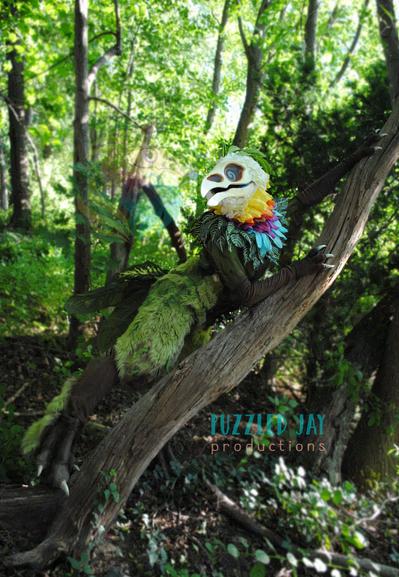 Costumed person dressed as a skeletal bird with colorful, floral plumage grabs a fallen tree in the forest and looks off into the distance. The same person in the same skeletal bird costume leans against an industrial support in a convention space. The bird creature looks up and over a fallen tree in the forest, but the picture is upside-down. Posed, indoor photograph against a gray backdrop showing the full, floral Curiosity costume.