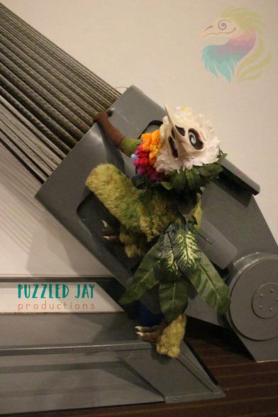 Costumed person dressed as a skeletal bird with colorful, floral plumage grabs a fallen tree in the forest and looks off into the distance. The same person in the same skeletal bird costume leans against an industrial support in a convention space. The bird creature looks up and over a fallen tree in the forest, but the picture is upside-down. Posed, indoor photograph against a gray backdrop showing the full, floral Curiosity costume.
