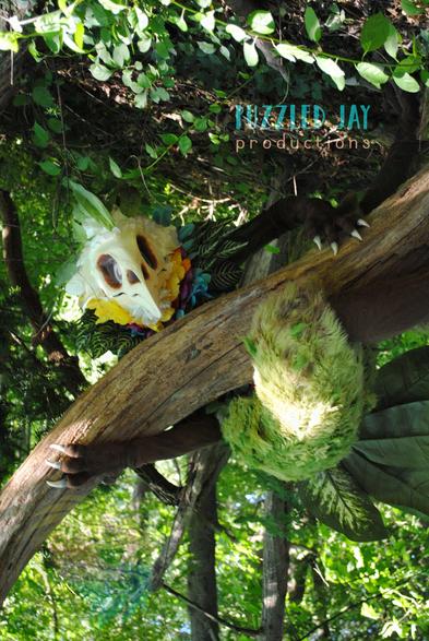Costumed person dressed as a skeletal bird with colorful, floral plumage grabs a fallen tree in the forest and looks off into the distance. The same person in the same skeletal bird costume leans against an industrial support in a convention space. The bird creature looks up and over a fallen tree in the forest, but the picture is upside-down. Posed, indoor photograph against a gray backdrop showing the full, floral Curiosity costume.