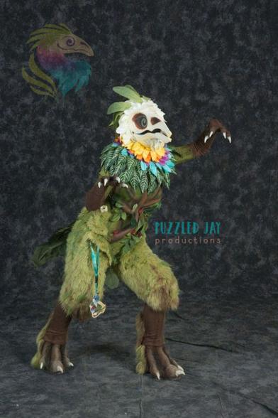 Costumed person dressed as a skeletal bird with colorful, floral plumage grabs a fallen tree in the forest and looks off into the distance. The same person in the same skeletal bird costume leans against an industrial support in a convention space. The bird creature looks up and over a fallen tree in the forest, but the picture is upside-down. Posed, indoor photograph against a gray backdrop showing the full, floral Curiosity costume.