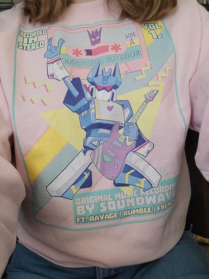 A photo of myself wearing a pink sweatshirt with art of the Transformers character Soundwave printed on it. The text and designs are evocative of a band shirt.