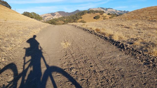 Photo of a brown, dirt road.  The shadow of a person astride a mountain bike is visible in the lower left of the photo.  The grass on both sides of the road and in the distant hills is golden-brown.  The blue sky with wisps of white clouds is visible at the top of the photo.