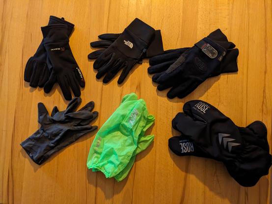 5 Handschuhe unterschiedlicher Dicke und 1 Handschuhüberzieher als Regenschutz auf einem Tisch.