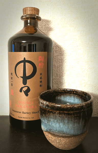 A bottle of Japanese shochu with a brown label. to the right is a ceramic cup with thick blue and black glaze on the upper half.