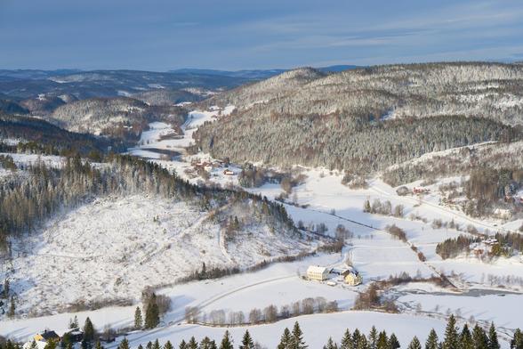 A snow covered landscape with open land in the foreground and forests in the background. The sky is cloudy, but still somewhat blue in colour