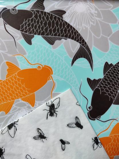 A tray whose design contains colourful carps. A napkin, white and with drawed flies, is on top, angular. It creates the rough illusion that carps are going to eat the flies.