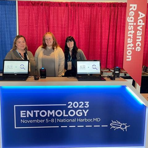 Three ESA staff members (Jess, Chloe, and Irene) stand behind a check-in counter with two laptop computers and barcode scanners. The lower front of the desk is royal blue with a white Entomology 2023 logo. Behind the staff is a tall red curtain and a sign that says "Advance Registration."