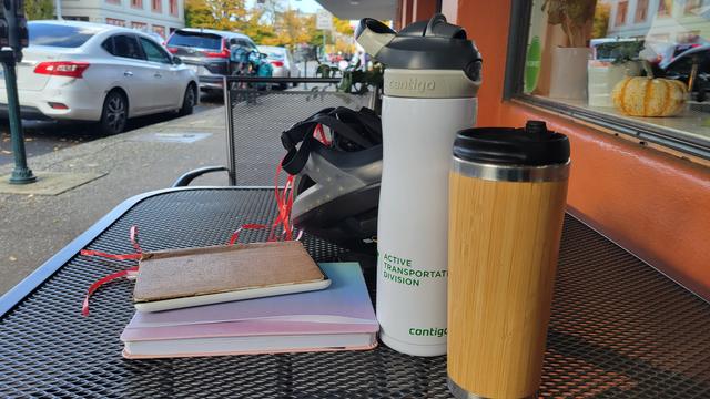 Photo of a black mesh wrought iron table against the orange wall of a building. On the table, a bamboo coffee travel cup, a white water bottle with the words Active Transportation Division printed on it in bright green, a couple of small books, and a black bike helmet with coral colored curling ribbon streamers. Beyond the table, a bicycle is locked to a nearby bike rack. The bike has a blue front bag and turquoise pannier.